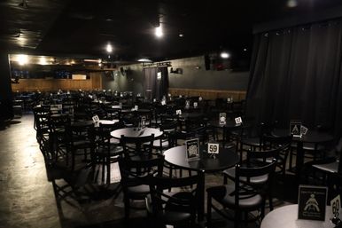 Dimly lit indoor comedy-club style venue with many round tables and black chairs, numbered table signs, and a small curtained stage ready for a show.