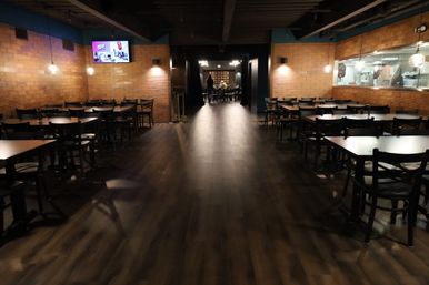 Spacious modern restaurant interior with dark wood floors, rows of empty black tables and chairs, exposed brick walls, hanging globe pendant lights, wall TV and a window into an open kitchen leading to a back dining room.