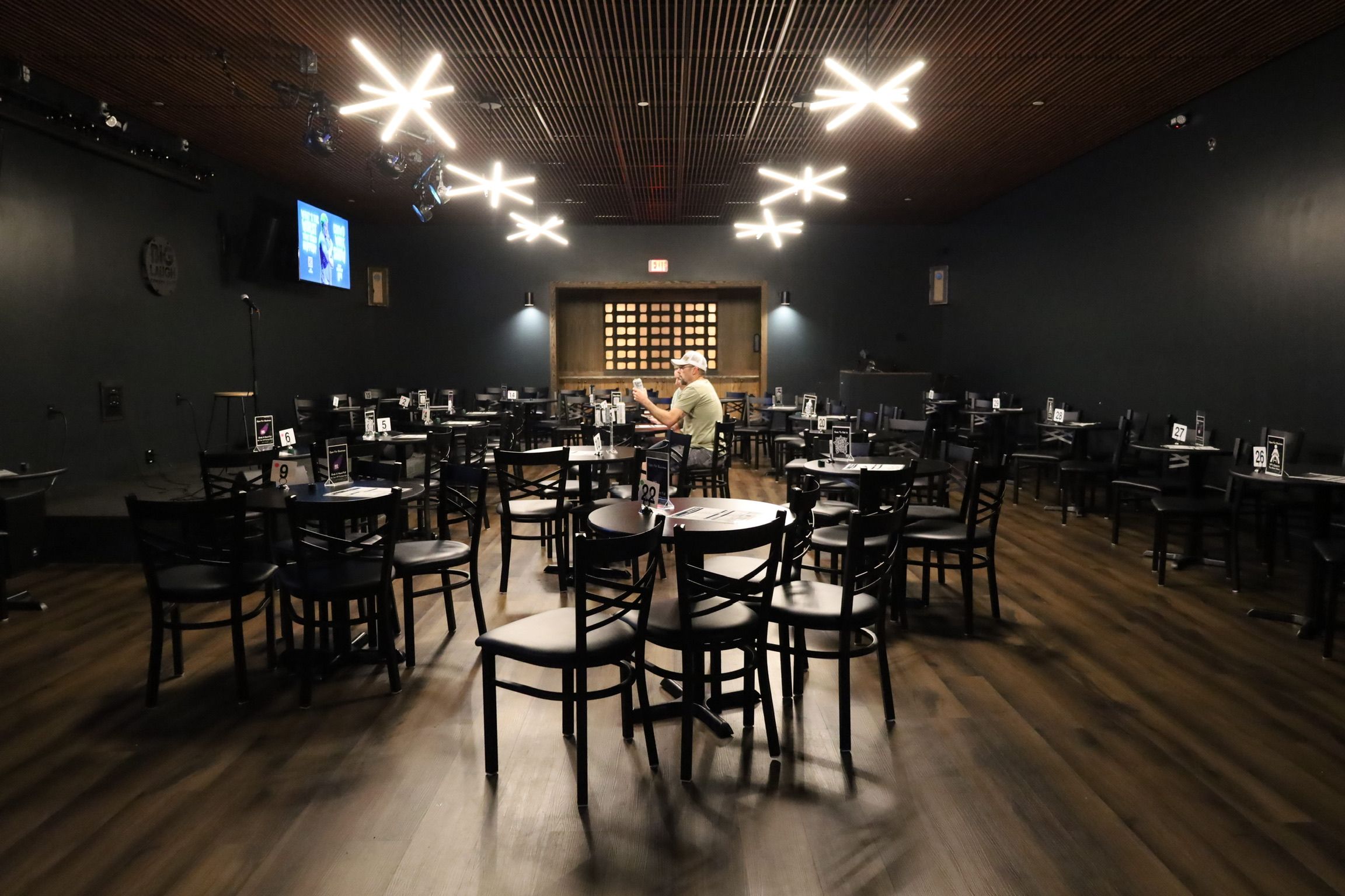 Nearly empty dimly lit event room with black chairs and round tables on a warm wood floor, a lone person seated center, small microphone-equipped stage to the left, a wall TV, and modern starburst ceiling lights overhead.