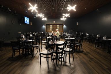 Nearly empty dimly lit event room with black chairs and round tables on a warm wood floor, a lone person seated center, small microphone-equipped stage to the left, a wall TV, and modern starburst ceiling lights overhead.