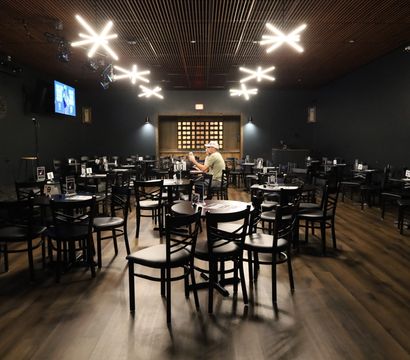 Nearly empty dimly lit event room with black chairs and round tables on a warm wood floor, a lone person seated center, small microphone-equipped stage to the left, a wall TV, and modern starburst ceiling lights overhead.