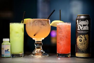 Colorful cocktails on a bar counter: a small green juice bottle, pale green iced drink with a straw, large goblet-style peach cocktail with a straw and citrus garnish, a tall red iced cocktail with a lemon wedge and straw, and a tall black can of sparkling beverage with bold white lettering.