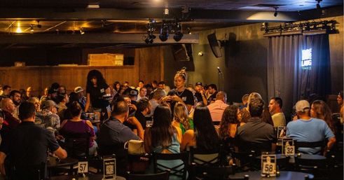 Crowded indoor comedy club scene with audiences seated at numbered tables, servers weaving through the crowd, a small stage with microphone and curtained backdrop under warm dim lighting for a lively night out