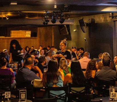 Crowded indoor comedy club scene with audiences seated at numbered tables, servers weaving through the crowd, a small stage with microphone and curtained backdrop under warm dim lighting for a lively night out