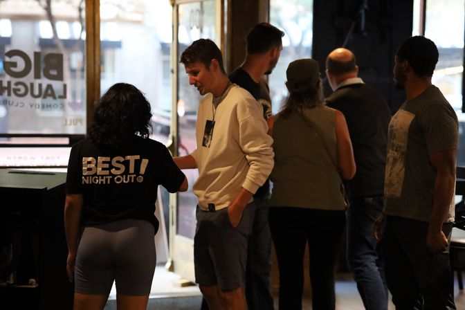 Casual group chatting near a dim venue entrance — a woman in a 'BEST! NIGHT OUT' tee talks with a man in a white sweatshirt while others queue to enter.
