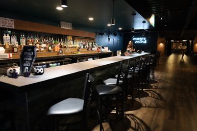 Cozy urban bar interior at night with a long concrete countertop, row of black bar stools, back shelves filled with liquor bottles, a glowing neon wall sign, bartender behind the bar, hardwood flooring, pendant lights and small Halloween decorations on the counter.