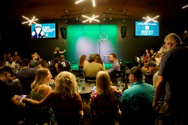 Crowded indoor comedy club with a green-lit stage and microphone, patrons seated at numbered tables with drinks and phones, ready for a live stand-up show — intimate nightlife venue.