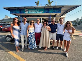 Eight adults in summer outfits smiling and posing in front of a blue pedal-powered party bike parked on a sunny beach-town street with palm trees and parked cars