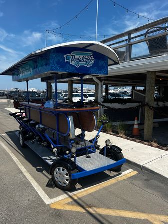 Blue multi-seat pedal-powered party bike with canopy parked in a sunny waterfront marina lot, boats and dockside boardwalk in the background