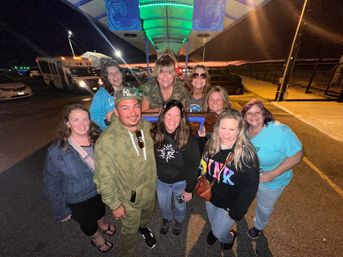 Group of friends smiling under a neon-green canopy in a nighttime outdoor parking lot, wearing casual jackets and hoodies for a fun night out.