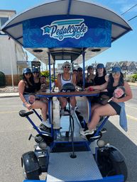 Smiling group of friends riding a blue pedal-powered party bike on a sunny coastal street lined with beach houses — summer group bike tour.