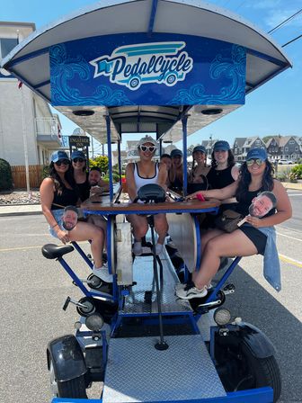 Smiling group of friends riding a blue pedal-powered party bike on a sunny coastal street lined with beach houses — summer group bike tour.