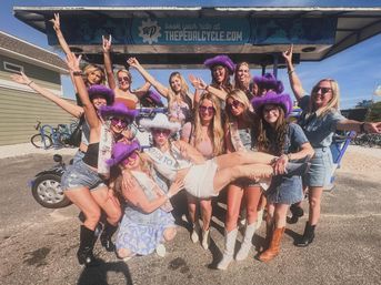 Group of women in purple cowboy hats and denim outfits cheerfully lift the bride-to-be wearing a sash and white dress in front of a sunlit beachside pedal-bar vehicle