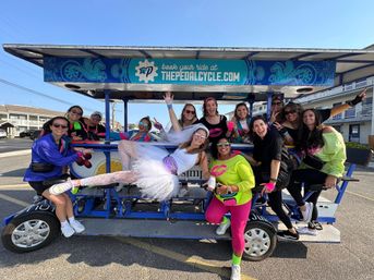 Cheerful group of friends in bright 80s-style outfits with a bride-to-be in a white tutu and veil riding a blue pedal-powered party bike on a sunny coastal street.