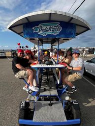 Smiling group riding a blue multi-person party pedal bike in a sunny waterfront parking lot near a marina, wearing colorful hats and ready to cruise.