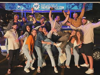 Group of friends cheering and posing at night around a brightly lit pedal pub party bike, casual denim outfits and big smiles in a lively street-party scene.