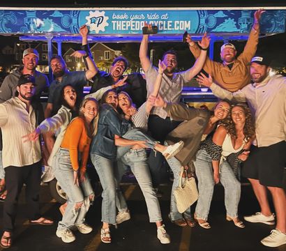Group of friends cheering and posing at night around a brightly lit pedal pub party bike, casual denim outfits and big smiles in a lively street-party scene.