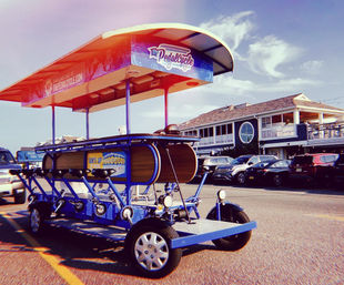Bright blue pedal pub (party bike) with canopy parked on a sunny beach‑town street in front of seaside restaurants and parked cars