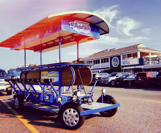 Bright blue pedal pub (party bike) with canopy parked on a sunny beach‑town street in front of seaside restaurants and parked cars