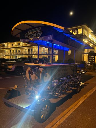 Night scene of a multi-seat pedal-powered party bike with blue LED accents and a bright headlight parked in a lit motel parking lot under a moonlit sky, evoking beach-town group tours.