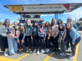 Group of smiling women in sunglasses posing in a sunny New Jersey parking lot under a sign that reads “Hey New Jersey, it’s that simple,” some holding playful face and dog photo cutouts with beach houses in the background.