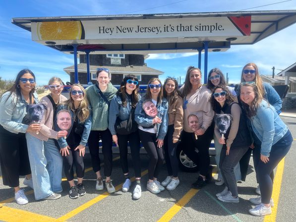 Group of smiling women in sunglasses posing in a sunny New Jersey parking lot under a sign that reads “Hey New Jersey, it’s that simple,” some holding playful face and dog photo cutouts with beach houses in the background.