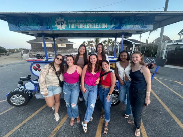 Smiling group of eight friends in summer outfits posing in front of a colorful multi-seat pedal party bike at a beachside parking lot with palm trees and sand dunes