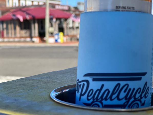 Close-up of a bright blue can cooler in a metal table cup holder at an outdoor cafe table, blurred small-town main street with a red awning and American flags in the background.