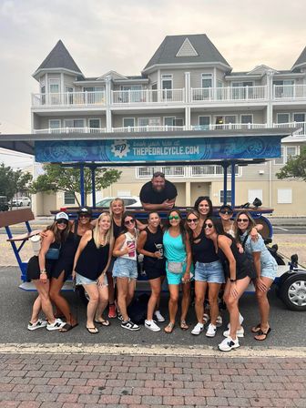 Smiling group of friends in summer outfits posing in front of a blue pedal-pub party bike on a coastal street with beachside condos behind them.