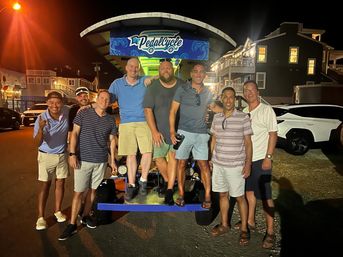 Group of eight men posing on and around a pedal-powered party bike at night on a lively coastal boardwalk street with illuminated shops and parked cars