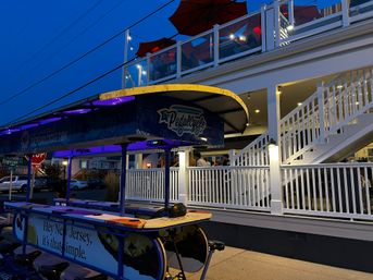Dusk scene of an illuminated pedal-party bike parked outside a two-story white restaurant with lit outdoor deck, red umbrellas and diners on the upstairs patio — lively evening atmosphere.
