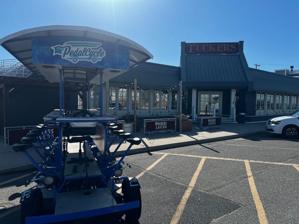 Blue pedal-powered party bike parked in a restaurant/tavern parking lot outside a gray-roofed tavern on a sunny day