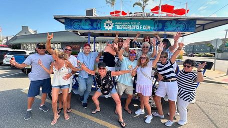 Group of adults smiling and waving aboard and around a blue party pedal-bike on a sunny beach-town street with red umbrellas, palm trees and storefronts in the background.