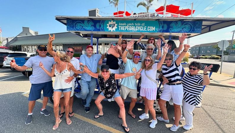 Group of adults smiling and waving aboard and around a blue party pedal-bike on a sunny beach-town street with red umbrellas, palm trees and storefronts in the background.