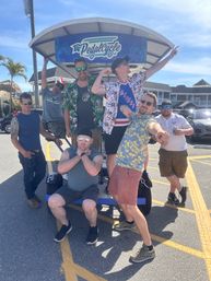 Seven friends laughing and striking playful poses on a blue pedal-bike party vehicle in a sunny coastal parking lot with palm trees and beach-style buildings, summer vacation vibe.
