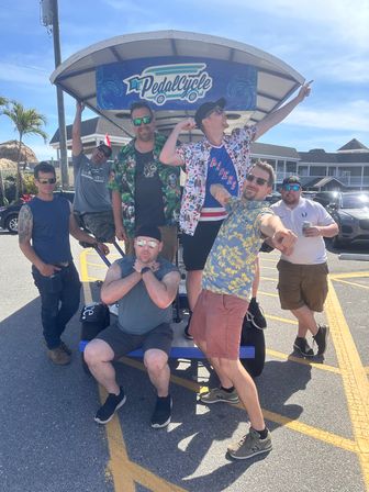 Seven friends laughing and striking playful poses on a blue pedal-bike party vehicle in a sunny coastal parking lot with palm trees and beach-style buildings, summer vacation vibe.