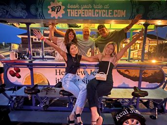 Five friends laughing and raising their arms on a colorful party pedal bike at a New Jersey boardwalk at dusk, string lights and beach shops glowing in the background