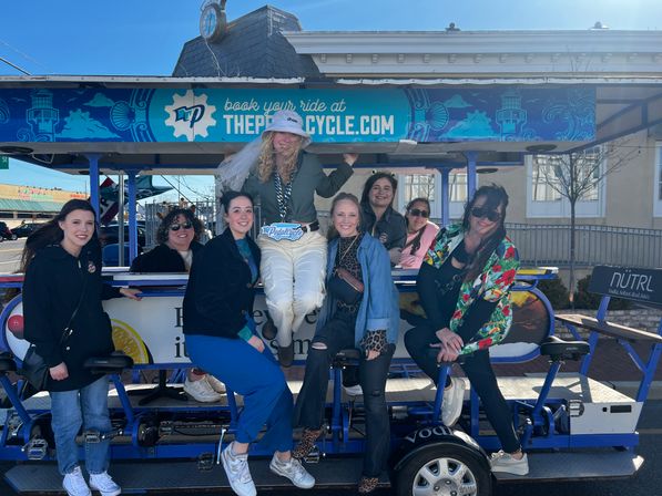 Eight women smiling on a blue outdoor pedal-pub bike on a sunny downtown street, bride-to-be wearing a veil and friends in casual colorful outfits for a bachelorette outing.