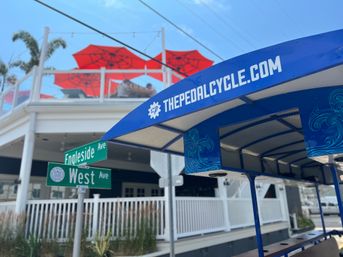 Blue covered party pedal bike parked at the corner of West Ave and Engleside Ave in front of a white-railed restaurant with bright red patio umbrellas on a sunny coastal day.