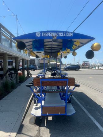 Sunlit blue pedal pub (party bike) parked on a waterfront street by a marina, long canopy with bench seating, pedals and steering wheel visible, black-and-gold balloons and boats in the background.