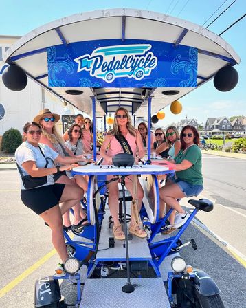 Smiling group of women on a blue pedal-powered party bike in a sunny coastal beach-town street, seated around a central bar table with balloons — summer pedal bike tour outing.