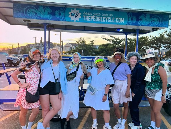 Smiling group of friends in summer outfits posing by a multi-seat pedal vehicle on a coastal boardwalk at sunset.