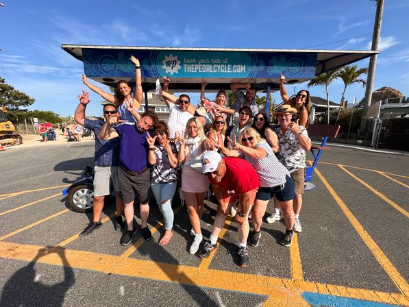 Group of adults cheering and posing on a colorful pedal-powered party bike in a sunny coastal parking lot with palm trees and blue sky.