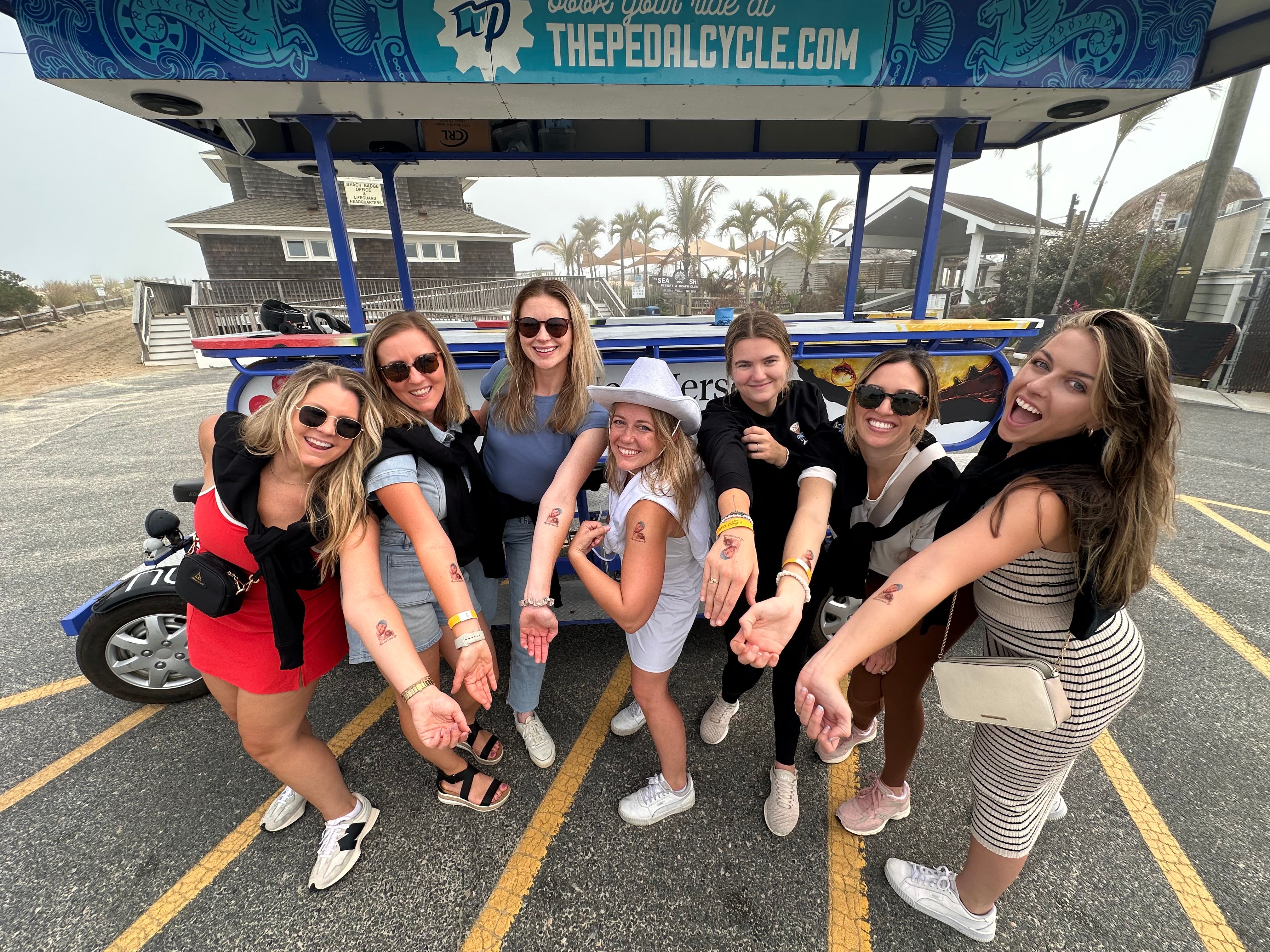 Eight women at a misty beach parking lot posing in front of a colorful pedal-bike tour vehicle, smiling and showing matching temporary tattoos on their forearms with palm trees and beach cottages behind them.