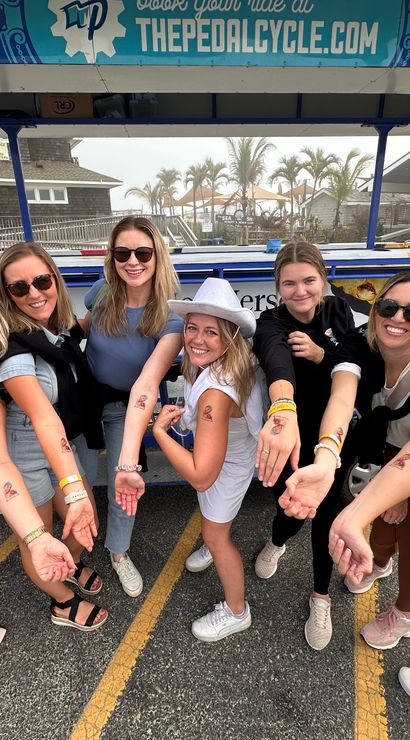 Eight women at a misty beach parking lot posing in front of a colorful pedal-bike tour vehicle, smiling and showing matching temporary tattoos on their forearms with palm trees and beach cottages behind them.