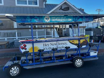 Blue multi-passenger pedal pub parked on a coastal New Jersey street — canopy-covered party bike with rows of saddles and pedals, citrus-and-berry ad panel, and a beach-style house and boardwalk railing behind it.