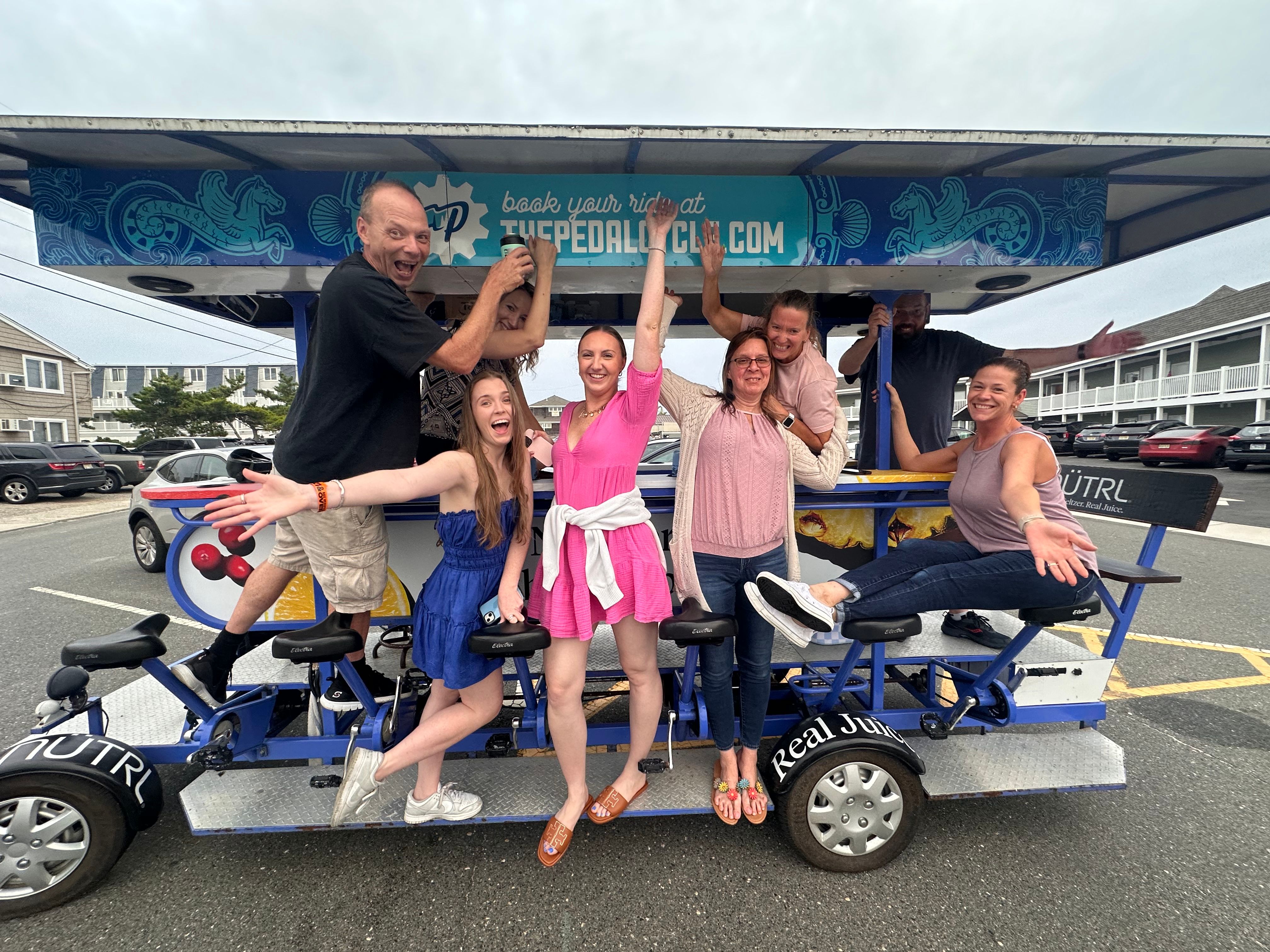 Eight adults cheering on a pedal-powered party bike in a coastal beach-town parking lot — lively summer group outing.