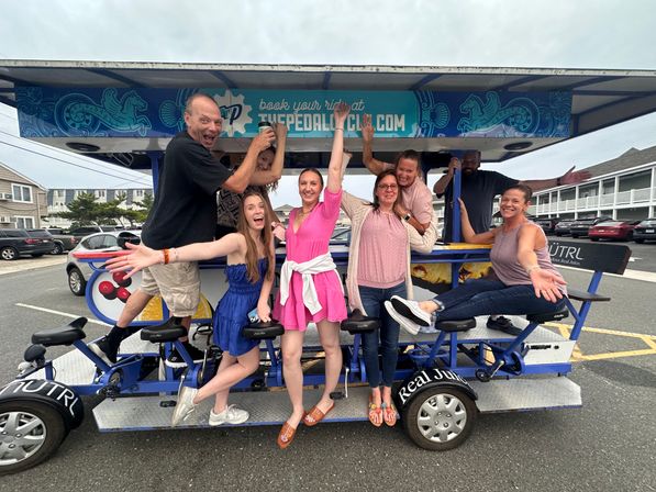Eight adults cheering on a pedal-powered party bike in a coastal beach-town parking lot — lively summer group outing.