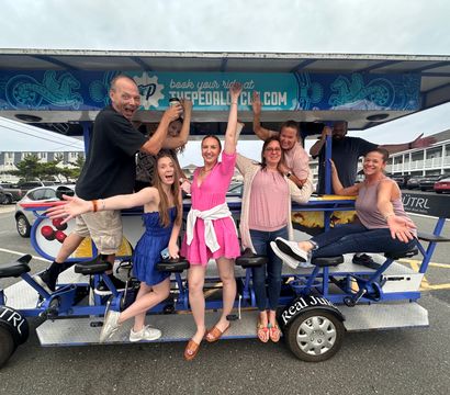 Eight adults cheering on a pedal-powered party bike in a coastal beach-town parking lot — lively summer group outing.