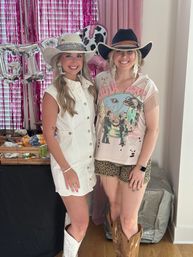 Two smiling women in cowgirl style outfits wearing cowboy hats and boots, one in a white button dress and embroidered hat, the other in a graphic tee with leopard shorts and a black hat, posing indoors in front of pink fringe backdrop and silver balloons.